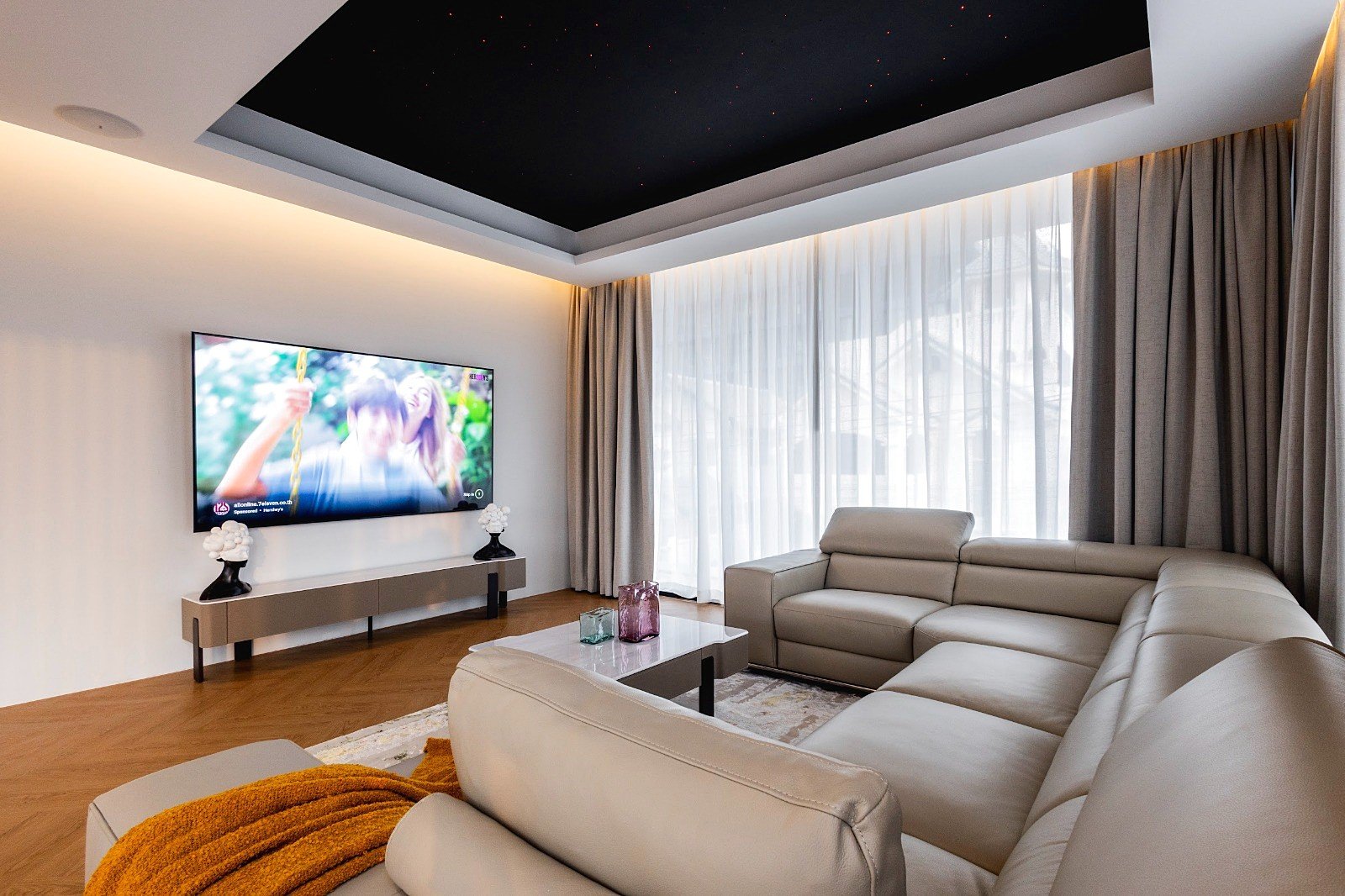 Casa_Jomtien_Village_Rent_Rdl352_38 A modern living room with an L-shaped beige leather sofa facing a wall-mounted flat-screen TV and a low console table with decorative lamps on either side.