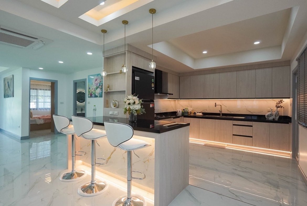 Open-plan modern kitchen with a light wood island, three white bar stools, marble floor, and under-cabinet lighting.