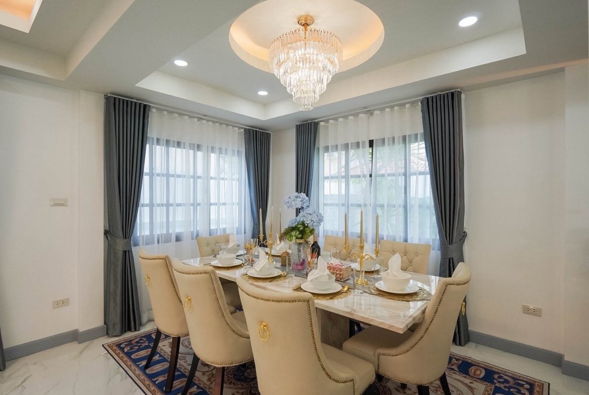 Elegant dining room with a marble table set for eight, gold candlesticks, and a crystal chandelier above; gray drapes and sheer curtains at large windows.