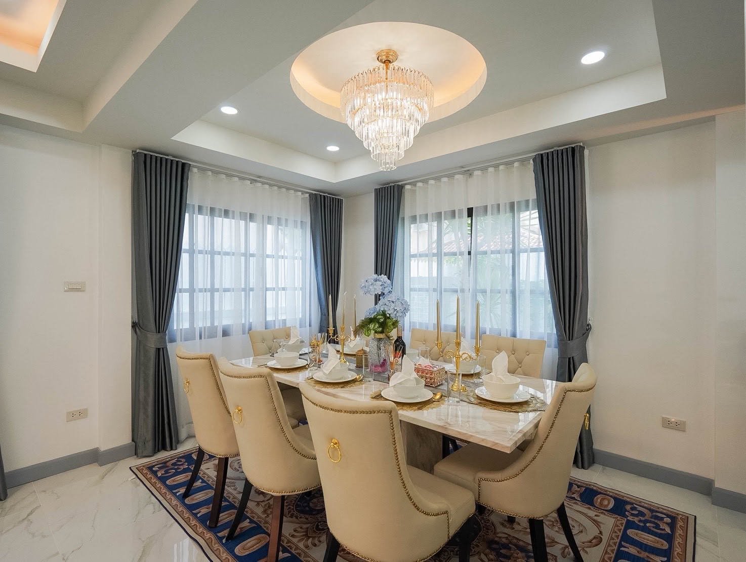 Central_Park_5_Pattaya_Sale_Dlh506_14 Elegant dining room with a marble table set for eight, gold candlesticks, and a crystal chandelier above; gray drapes and sheer curtains at large windows.