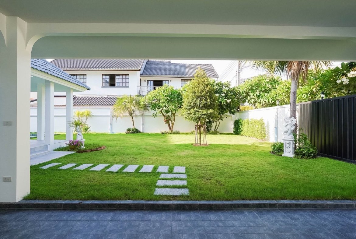Backyard scene with a covered patio, a green lawn, and stepping-stone path leading toward trees and a decorative statue beside a white fence.