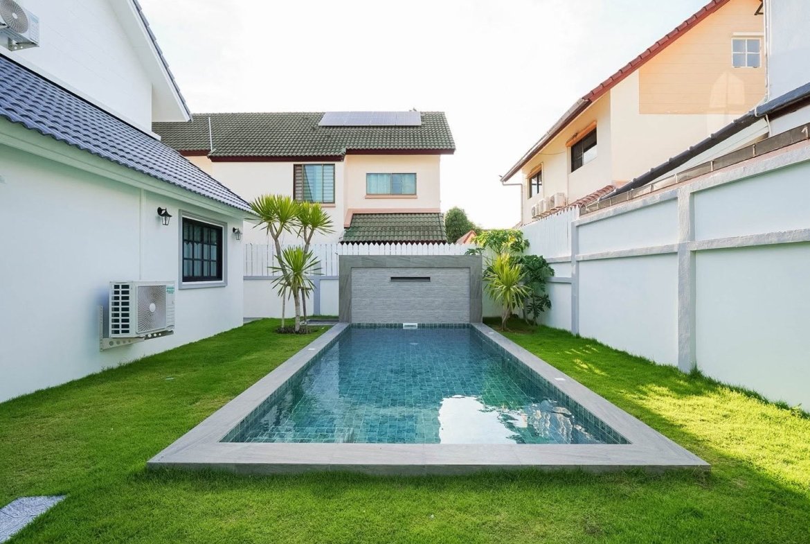 Long rectangular swimming pool in a tidy backyard, flanked by white walls, green grass, and small palm trees.