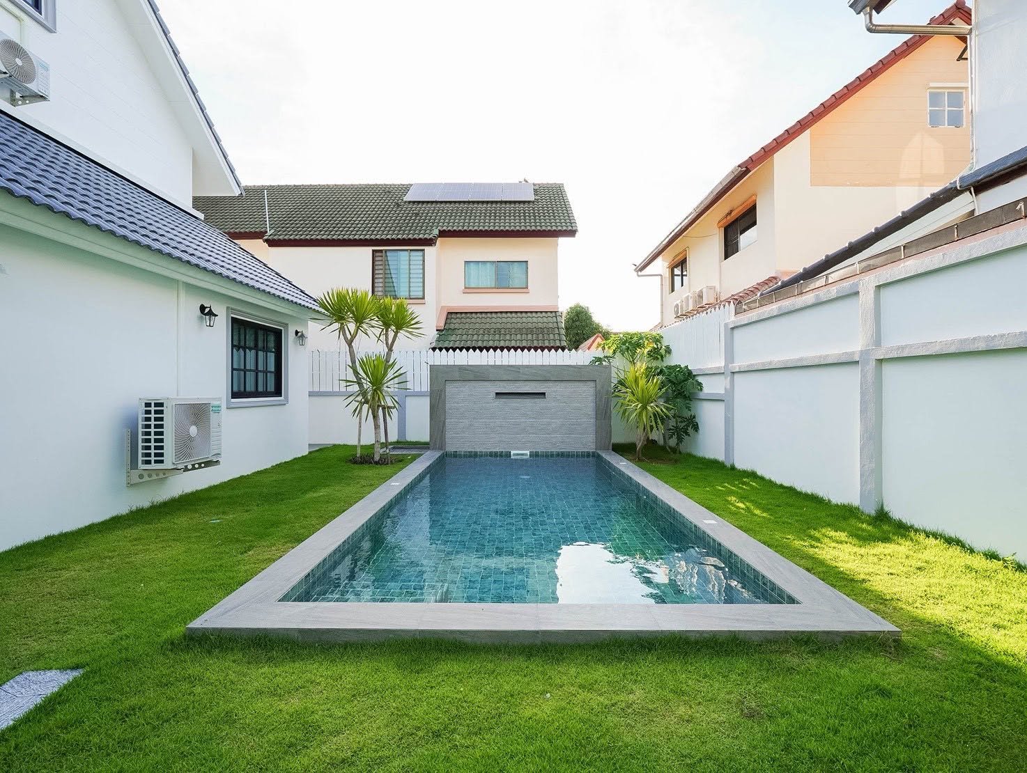 Central_Park_5_Pattaya_Sale_Dlh506_2 Long rectangular swimming pool in a tidy backyard, flanked by white walls, green grass, and small palm trees.