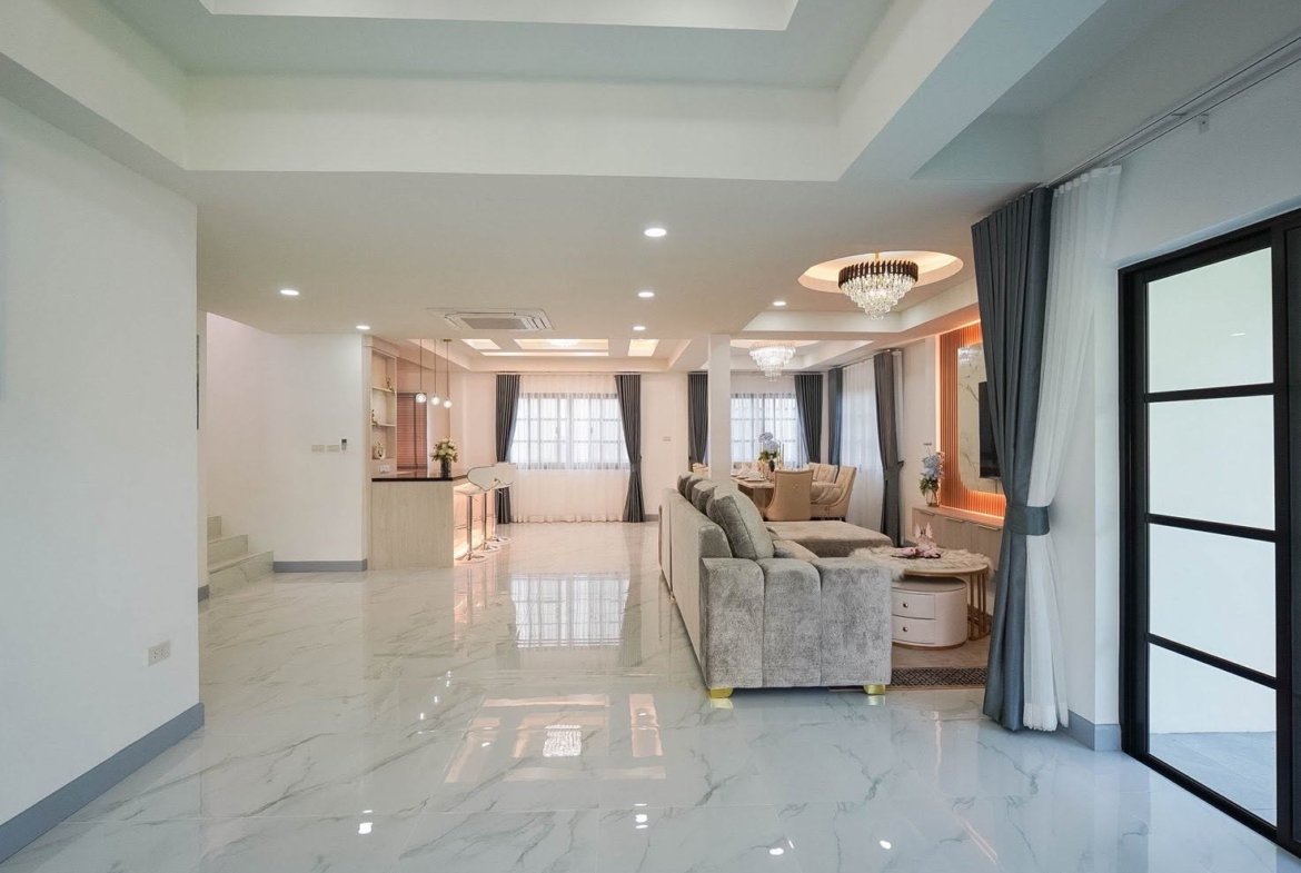 Open-plan living room with a marble floor, gray sectional sofa, and a dining area in the background under chandeliers.