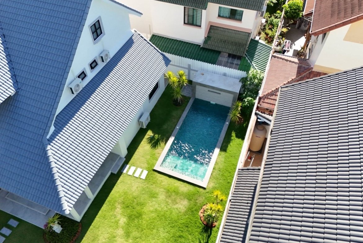 Aerial view of a modern backyard with a rectangular in-ground pool, lush green lawn, and palm trees, surrounded by white fencing and blue-gray tiled roofs.