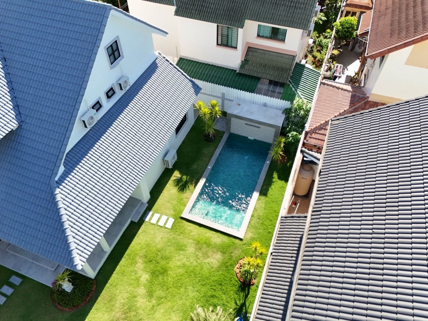 Central_Park_5_Pattaya_Sale_Dlh506_23 Aerial view of a modern backyard with a rectangular in-ground pool, lush green lawn, and palm trees, surrounded by white fencing and blue-gray tiled roofs.