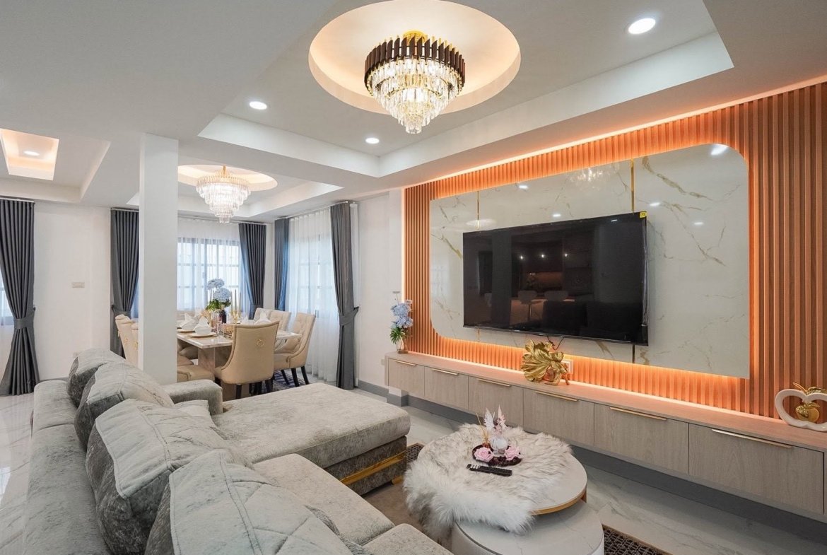 Luxurious living room with an L-shaped gray sofa facing a wall-mounted TV and marble backdrop under warm lighting.