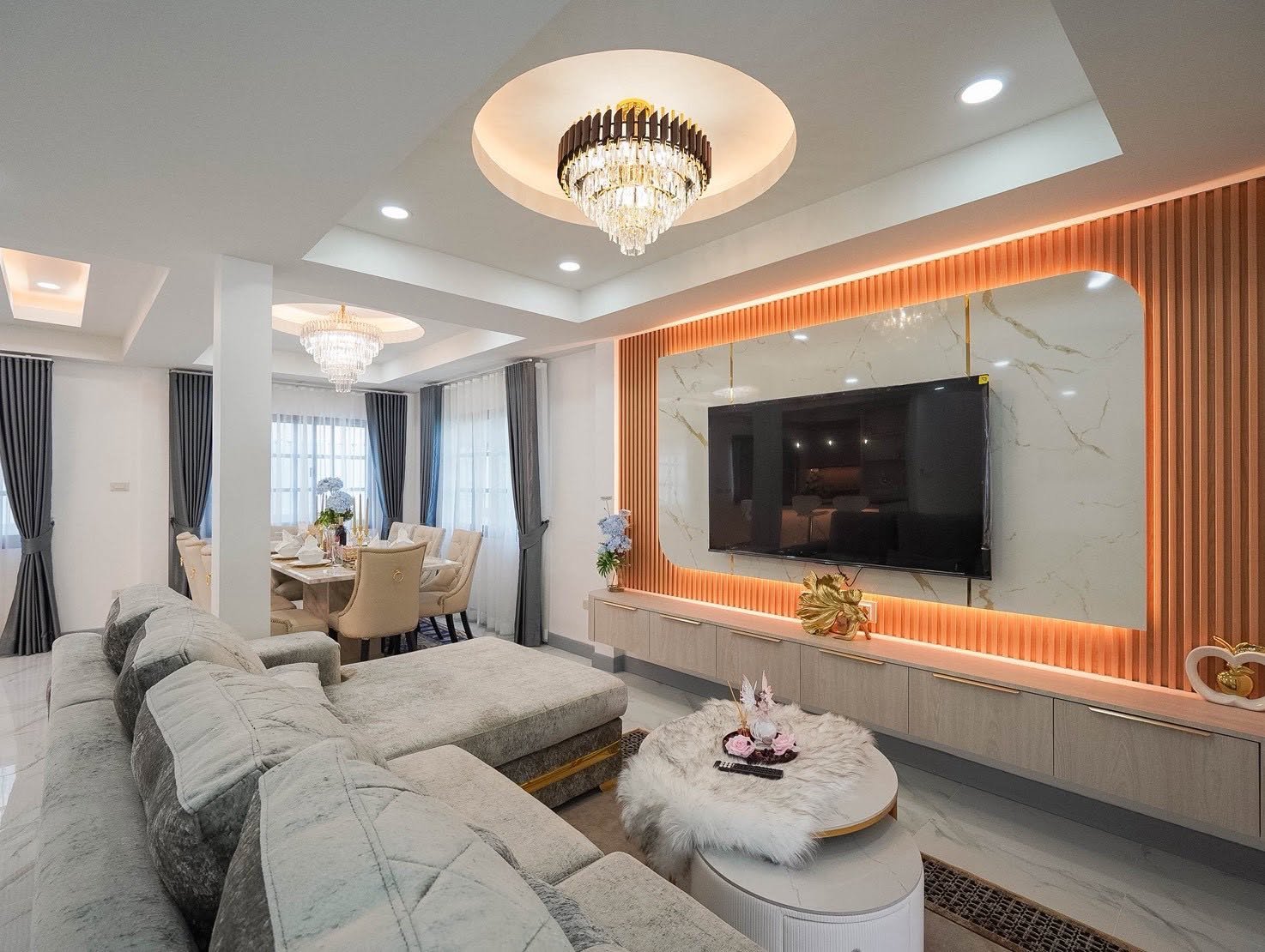 Central_Park_5_Pattaya_Sale_Dlh506_3 Luxurious living room with an L-shaped gray sofa facing a wall-mounted TV and marble backdrop under warm lighting.