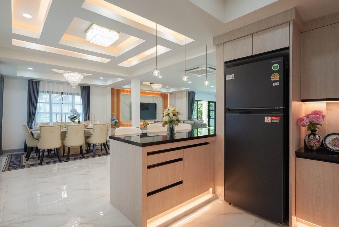 Open-concept dining and kitchen area with a light wood island, a large black refrigerator to the right, and a marble floor; a formal dining table with cream chairs is visible in the background under crystal chandeliers.
