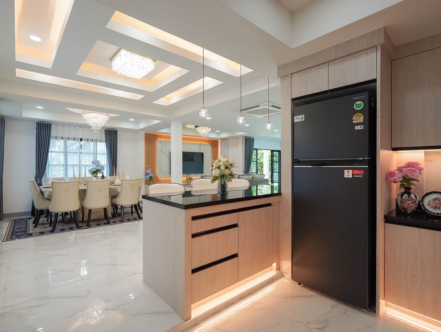 Central_Park_5_Pattaya_Sale_Dlh506_4 Open-concept dining and kitchen area with a light wood island, a large black refrigerator to the right, and a marble floor; a formal dining table with cream chairs is visible in the background under crystal chandeliers.