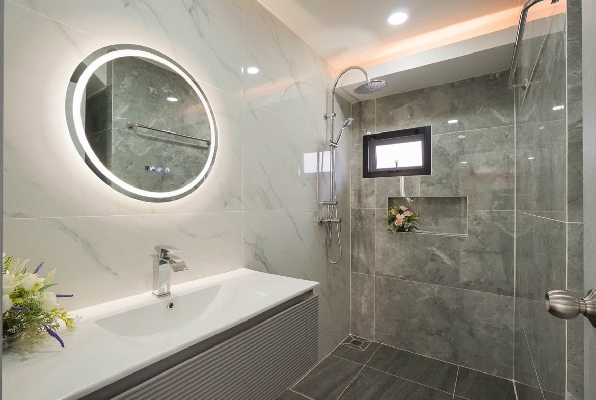 Modern bathroom with a round backlit mirror above a white sink, gray marble walls, and a glass shower with a rain head and built-in niche flowers.