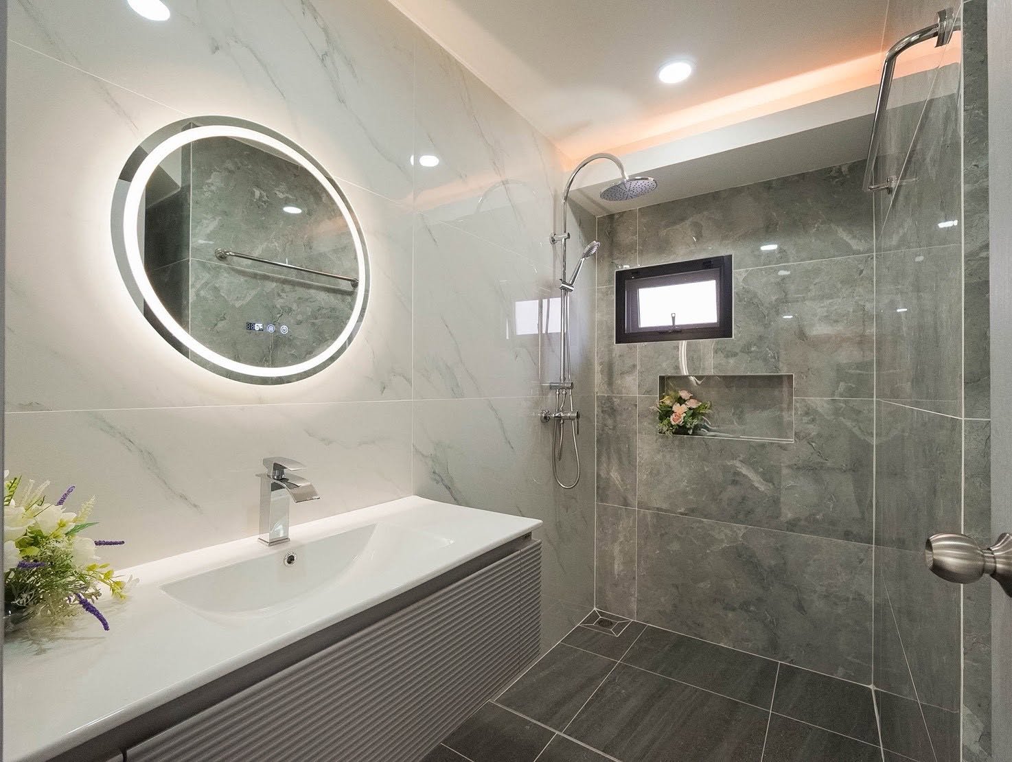 Central_Park_5_Pattaya_Sale_Dlh506_7 Modern bathroom with a round backlit mirror above a white sink, gray marble walls, and a glass shower with a rain head and built-in niche flowers.