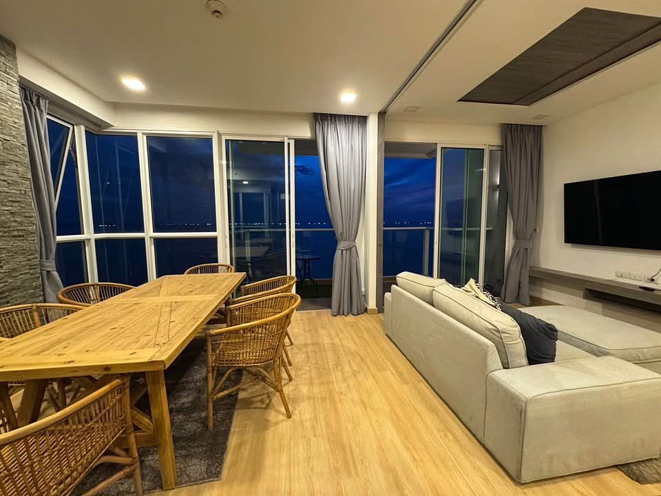Open-plan living and dining room with a long wooden dining table and woven chairs, a beige sectional sofa, and a wall-mounted TV, overlooking a balcony at night.