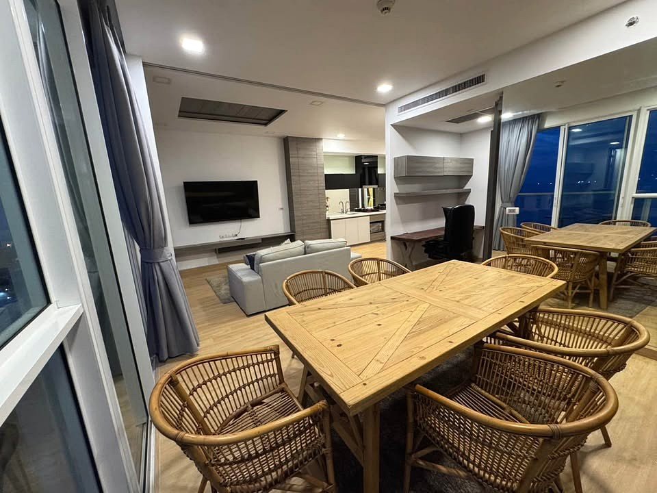 Open-concept living/dining area with a wooden table and wicker chairs, gray sofa, and a wall-mounted TV. Behind it is a small kitchen.