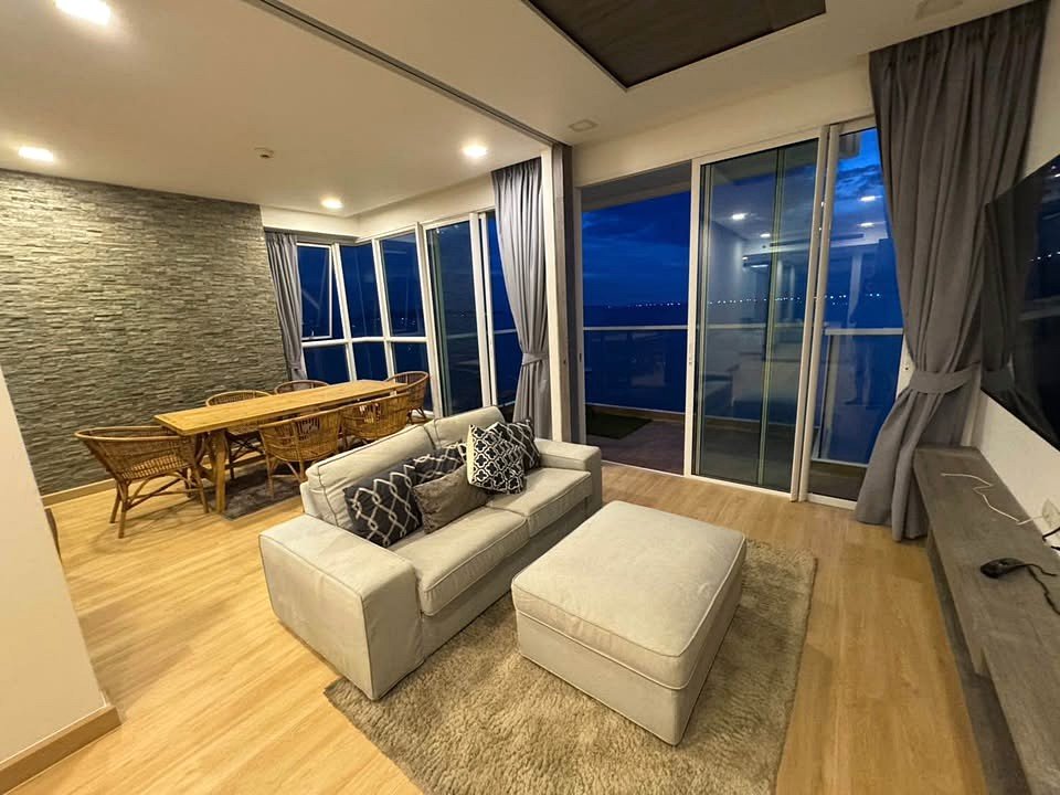 Open-plan living and dining area: gray sofa with ottoman, wooden dining table and wicker chairs, stone accent wall, and floor-to-ceiling windows with gray curtains at night.