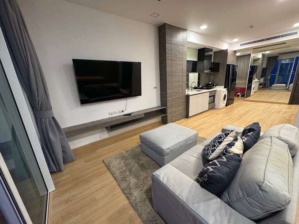 Open-plan living area with a wall-mounted TV, gray sectional sofa, and built-in shelf; kitchen and appliances visible in the background.