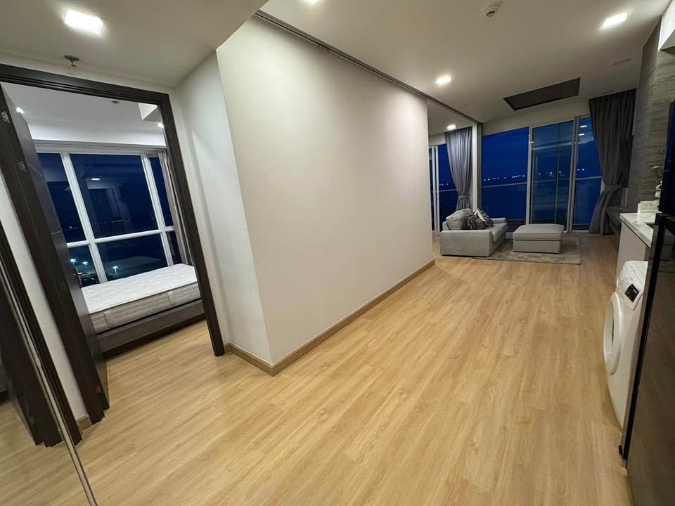 Open living area at night with large windows showing city lights, gray sofa and ottoman, light wood floors, and a washing machine near the kitchen.
