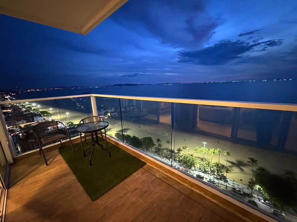 Nighttime balcony overlooking a calm sea with a glass railing, small table and chairs on a wooden deck, palm trees below.