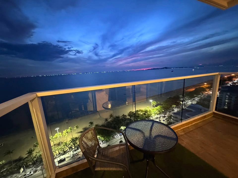 Ocean-front balcony at dusk with glass railing, table and chair, overlooking palm-lined street and distant lights.
