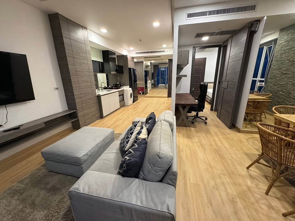 Open-plan living area with a gray sectional sofa facing a wall TV, light wood floors, and a modern kitchen in the background.