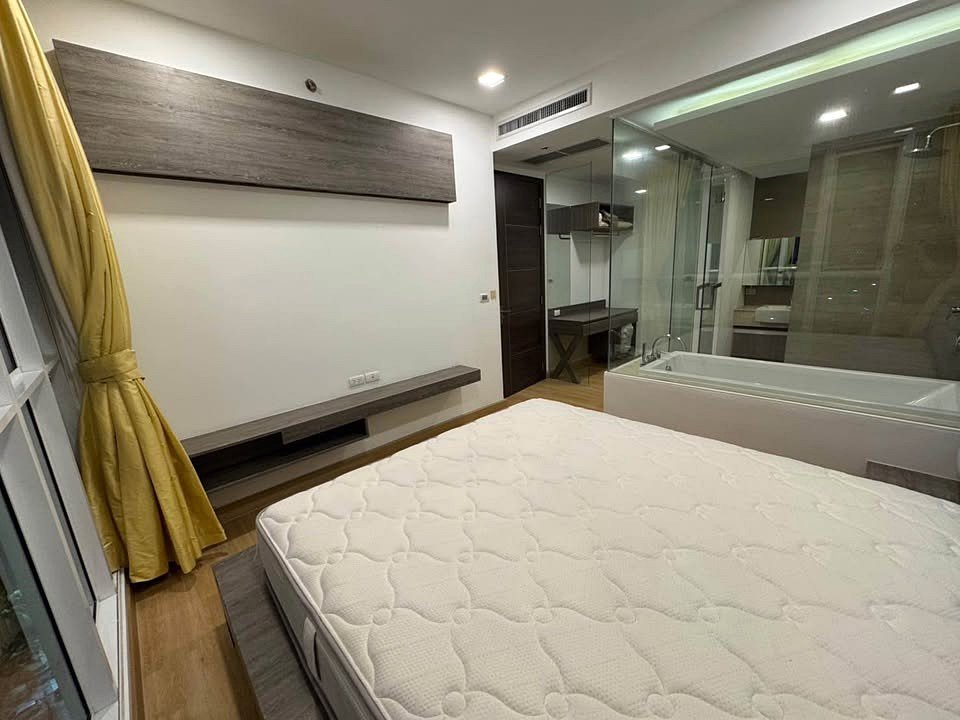 Modern hotel room with a platform bed, wall-mounted shelf, and a glass-enclosed bathroom visible to the right; yellow curtains at a window.