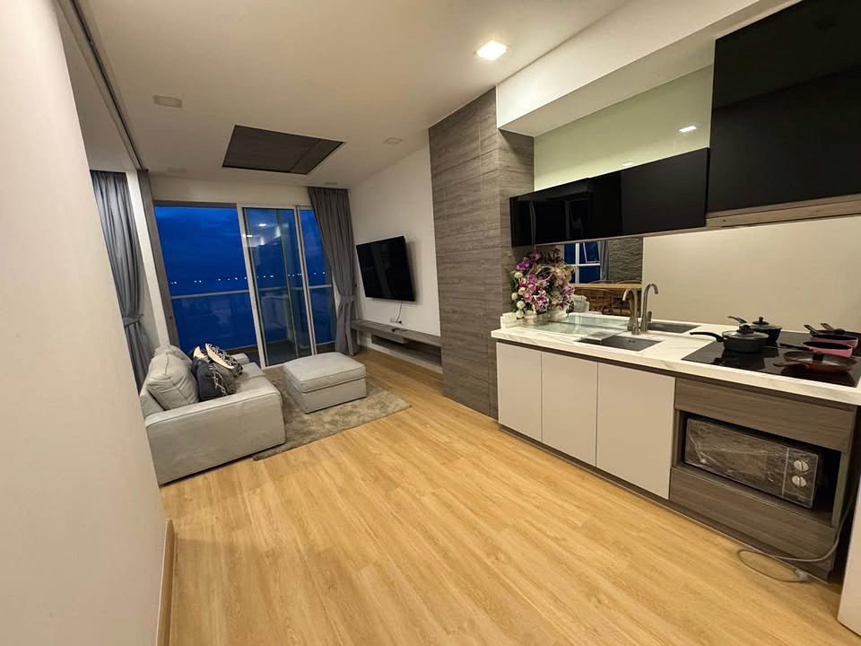 Open-plan living area with a gray sofa, ottoman, and a wall-mounted TV, opening to a balcony with blue evening light. A modern kitchen with white cabinets and a flower bouquet on the counter sits to the right.