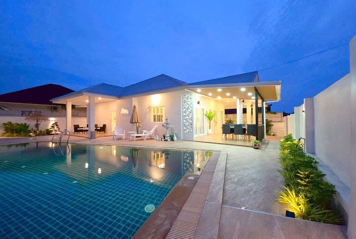 Modern backyard at dusk: illuminated pool with blue-tiled water and surrounding lounge area.