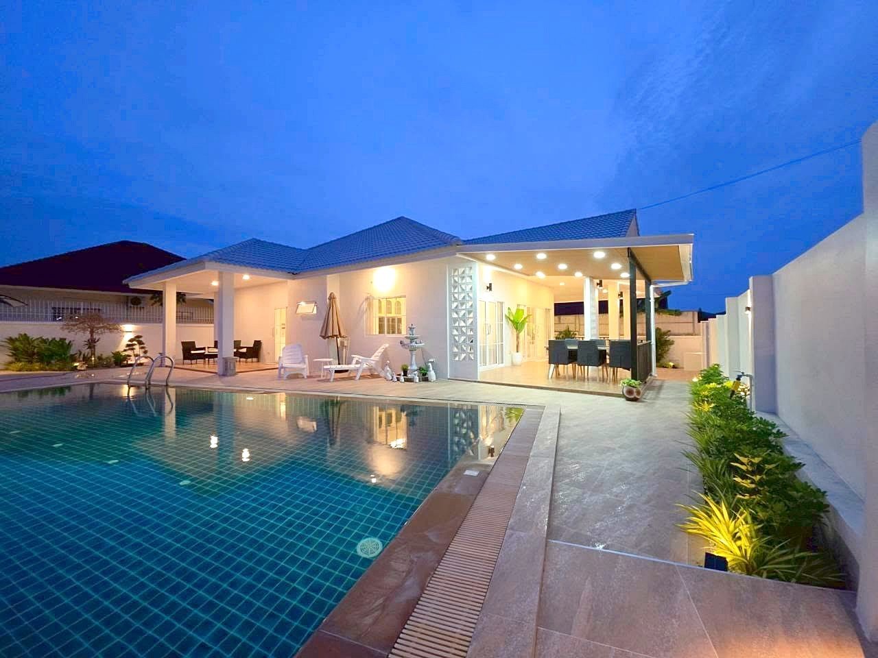 Modern backyard at dusk: illuminated pool with blue-tiled water and surrounding lounge area.