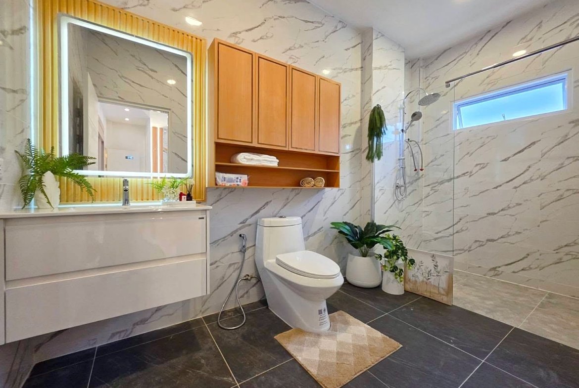 Modern bathroom with marble walls, wooden cabinets, and a glass shower; toilet, vanity with illuminated mirror, and potted plants.