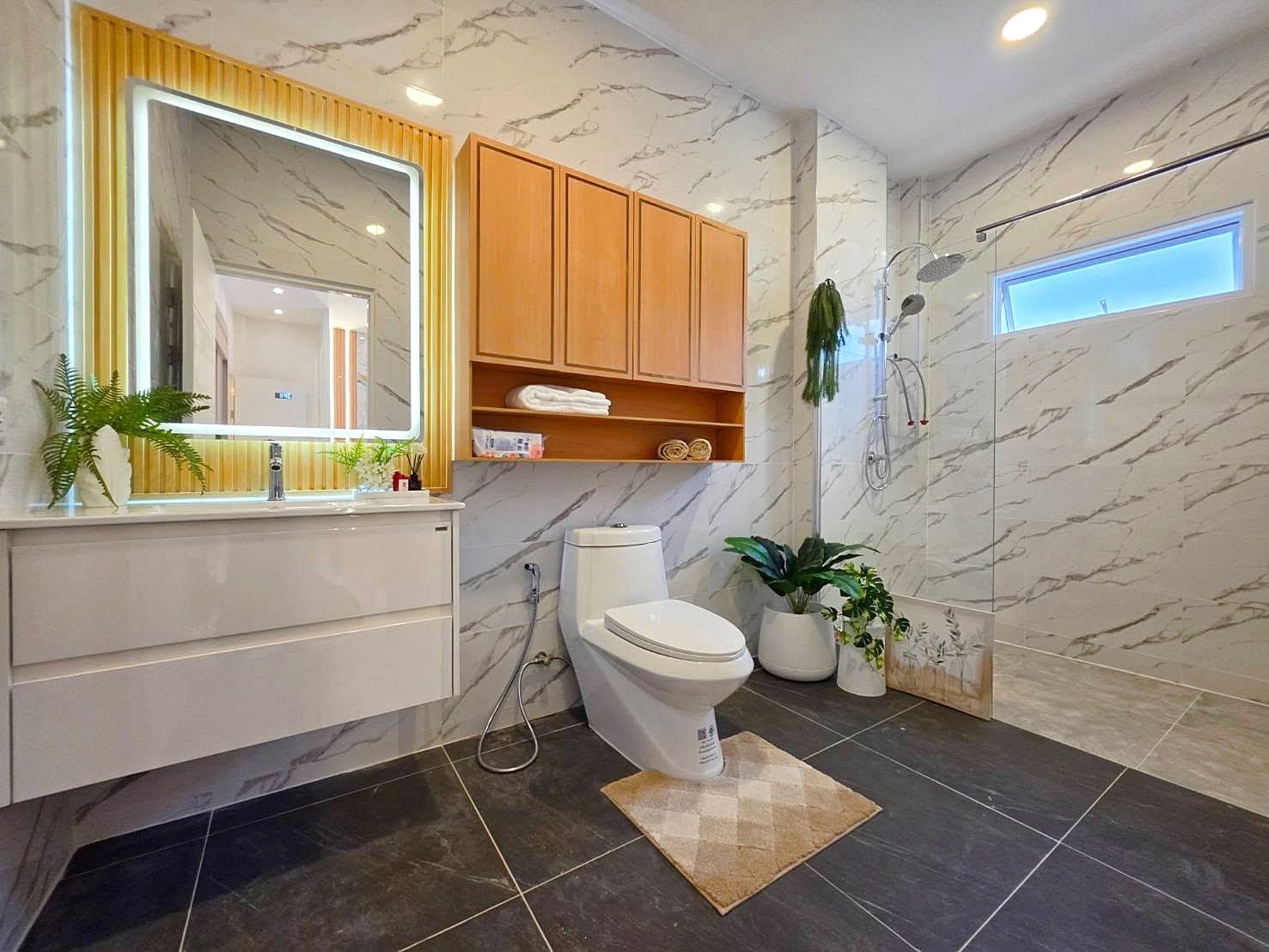 Modern bathroom with marble walls, wooden cabinets, and a glass shower; toilet, vanity with illuminated mirror, and potted plants.