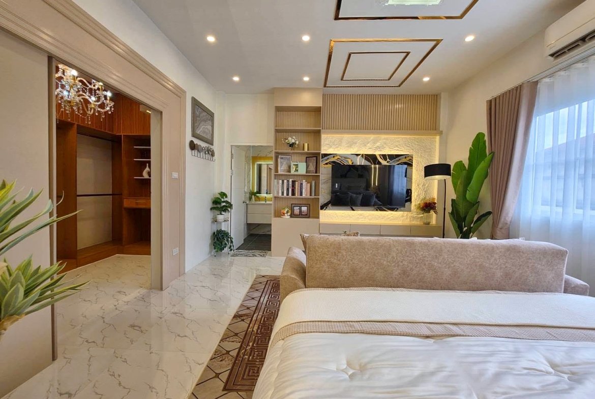 Luxurious bedroom with a beige bed and seating area facing a wall-mounted TV in a softly lit living space.