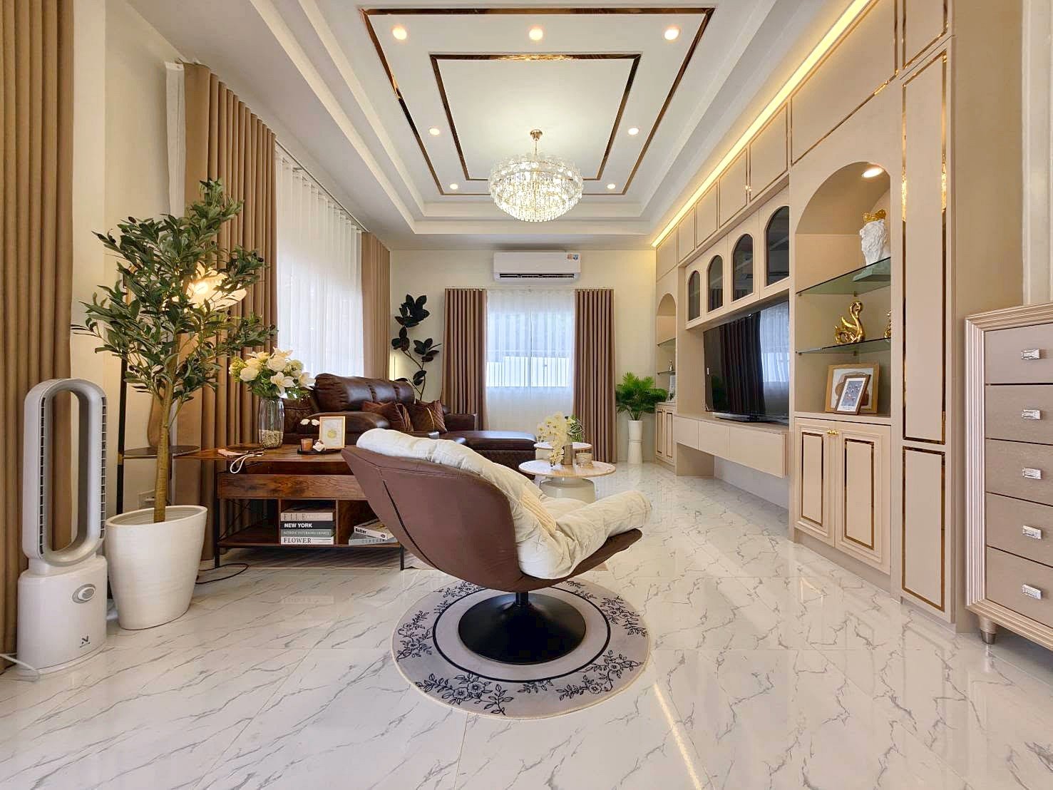 Bright living room with marble floor, beige curtains, brown sofa, swivel chair, and a built-in entertainment wall with a chandelier overhead.