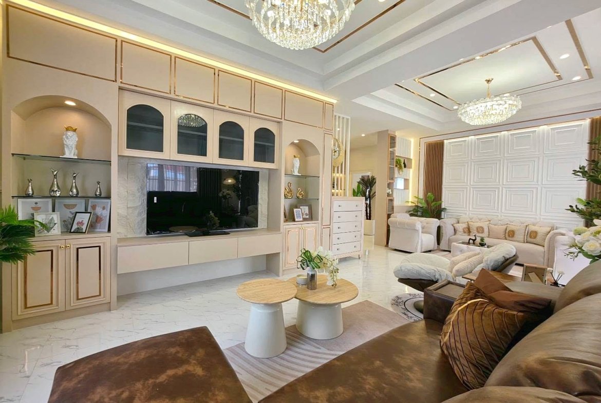 Luxurious living room with a built-in beige TV wall unit, crystal chandelier, and white marble flooring, cream sofas, and greenery.