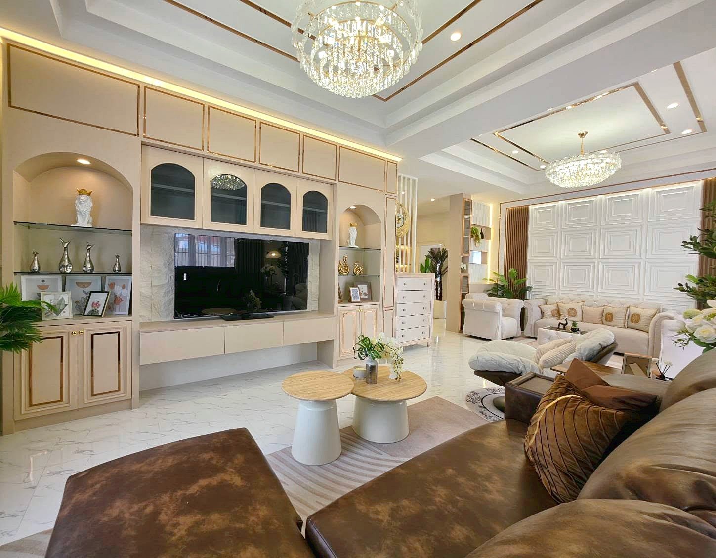 Luxurious living room with a built-in beige TV wall unit, crystal chandelier, and white marble flooring, cream sofas, and greenery.