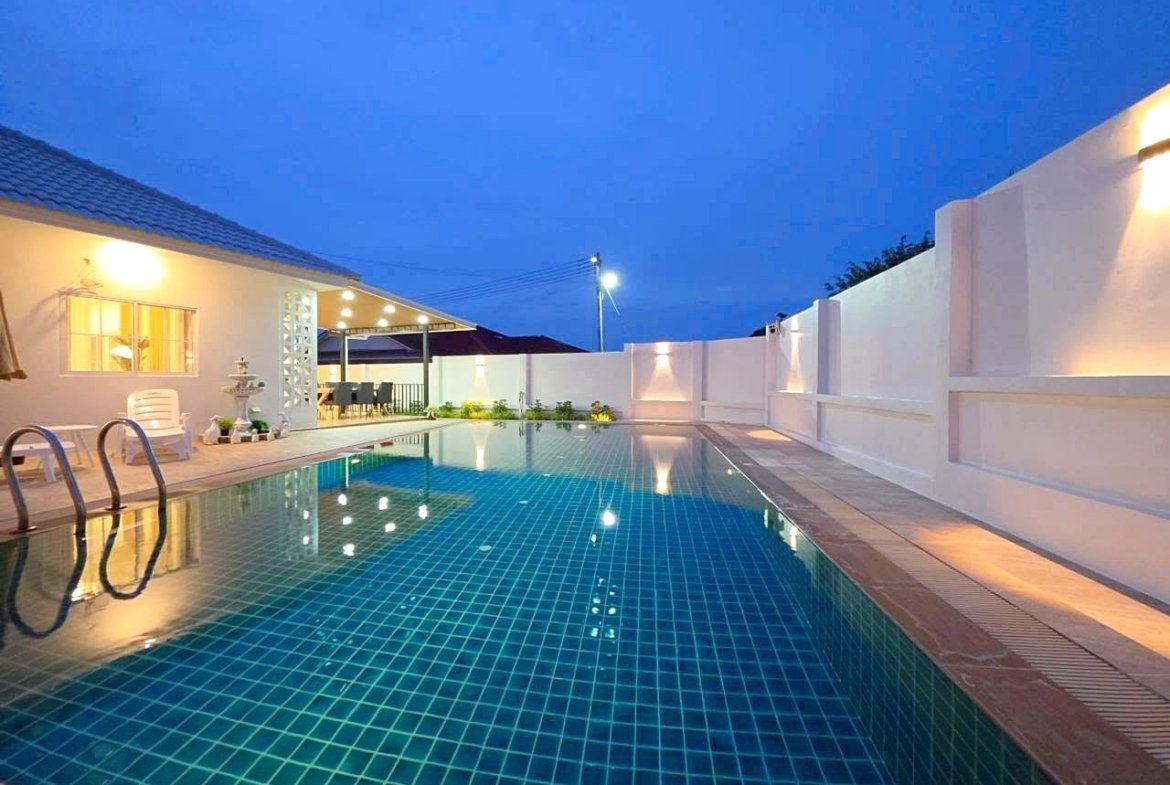 Evening view of a rectangular backyard pool with blue tiled water, white walls, and warm patio lights.