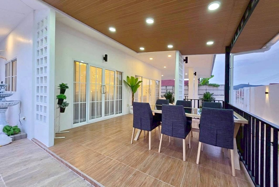 Outdoor covered patio with a long dining table and eight dark chairs, glass doors, and potted plants.