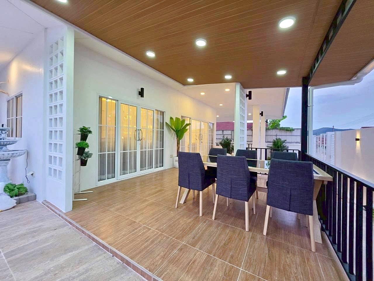 Outdoor covered patio with a long dining table and eight dark chairs, glass doors, and potted plants.