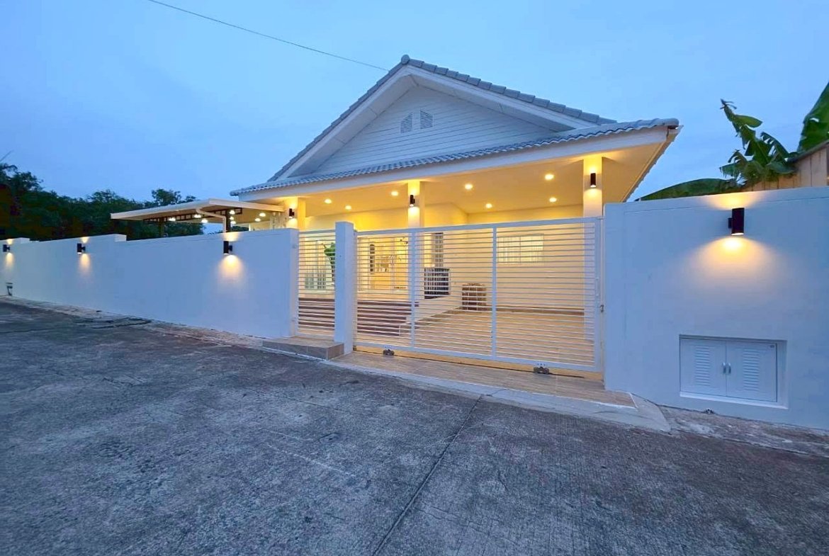 White modern house with a sloped roof, illuminated entrance, and white horizontal-slatted gate at dusk/early evening.