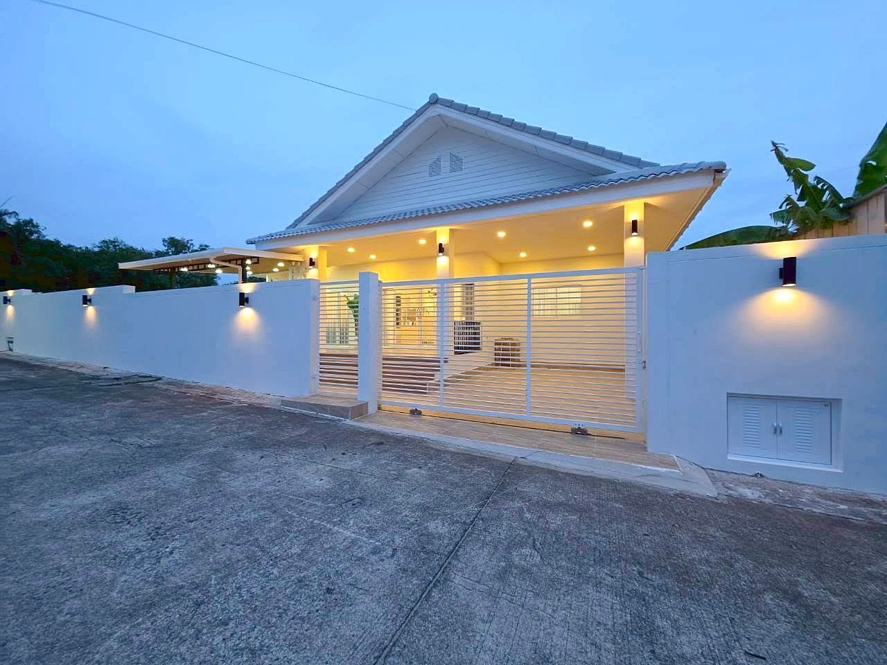 White modern house with a sloped roof, illuminated entrance, and white horizontal-slatted gate at dusk/early evening.