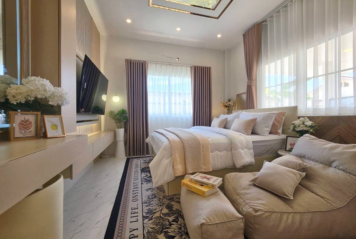 Bright modern bedroom with a large bed in white and beige bedding, a beige sofa, and sheer white curtains by floor-to-ceiling windows, plus plants and art on the dresser.