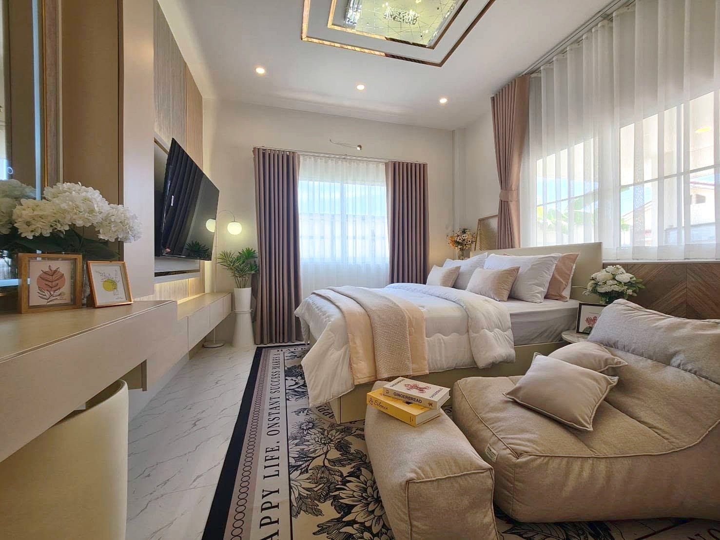 Bright modern bedroom with a large bed in white and beige bedding, a beige sofa, and sheer white curtains by floor-to-ceiling windows, plus plants and art on the dresser.