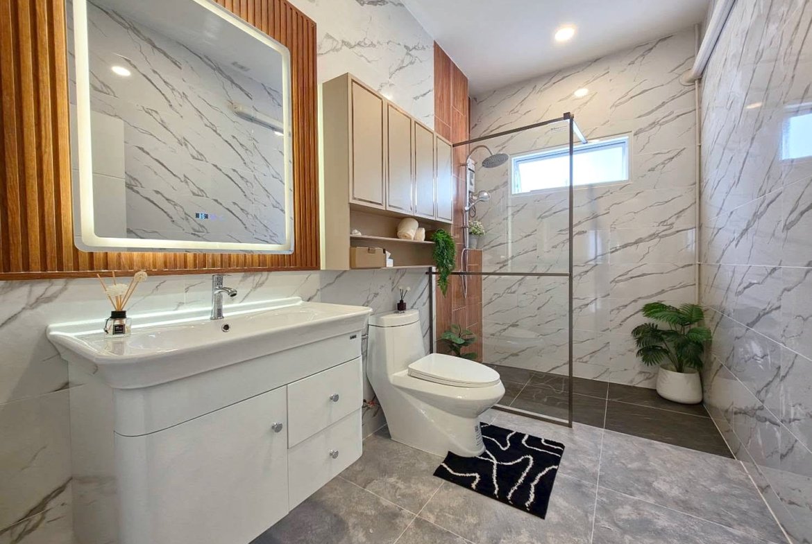 Modern bathroom with marble walls, white vanity, toilet, and glass-enclosed shower; potted plants add greenery.