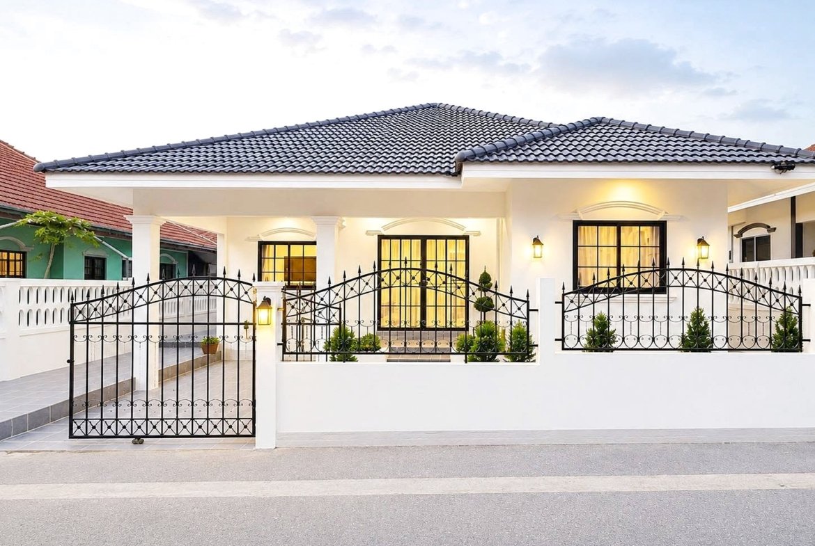 White, single-story house with black wrought-iron gate and decorative fence, illuminated entrance and windows, potted plants along the front