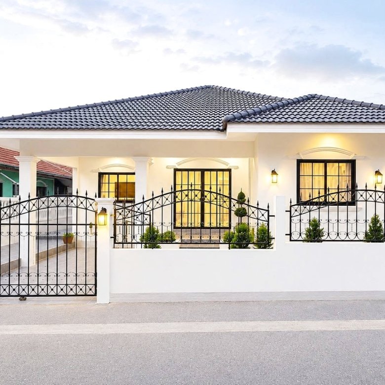 White single story house with black wrought iron gate and decorative fence illuminated entrance and windows potted plants along the front