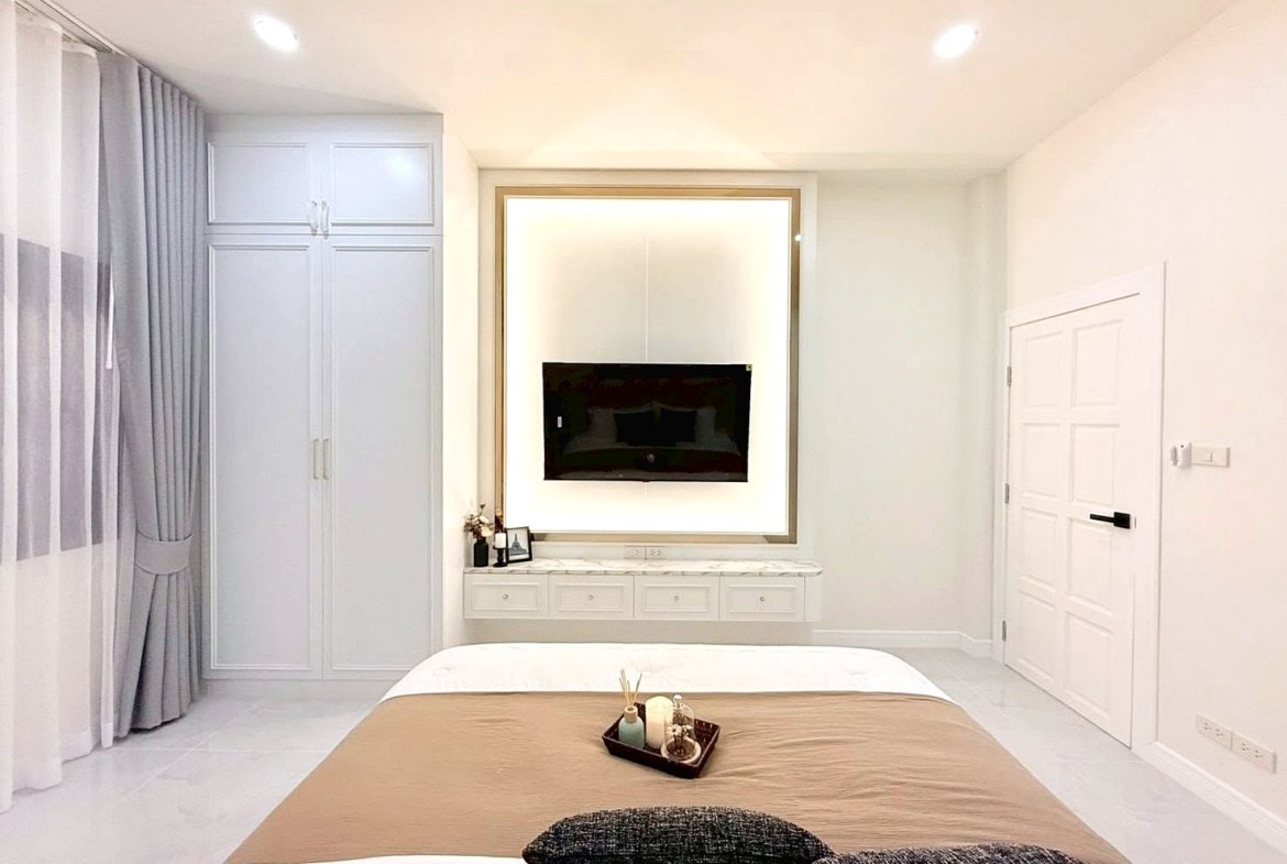 Minimalist bedroom with beige bedspread, white furniture, and a wall-mounted TV in a backlit frame.