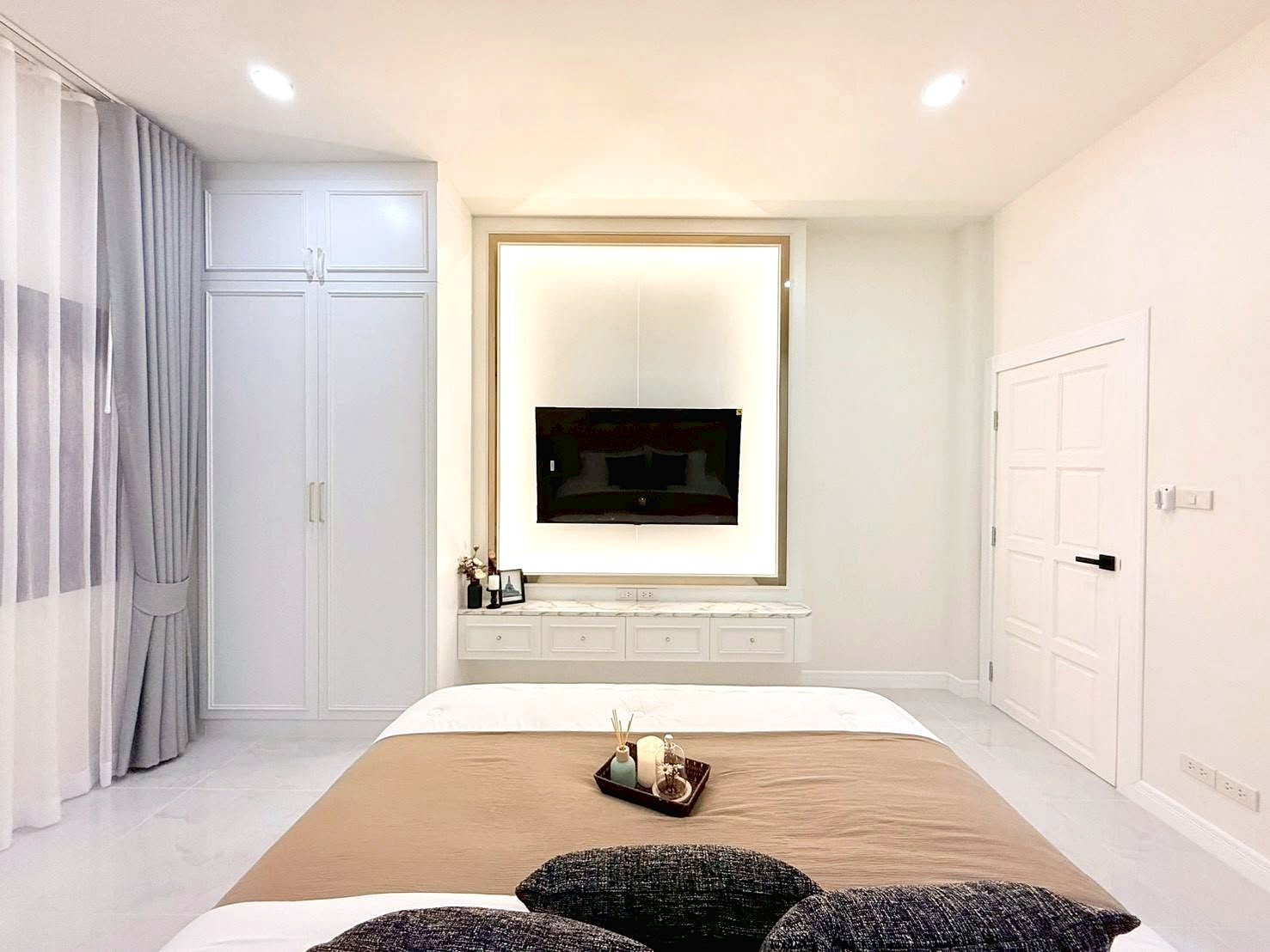 Minimalist bedroom with beige bedspread, white furniture, and a wall-mounted TV in a backlit frame.