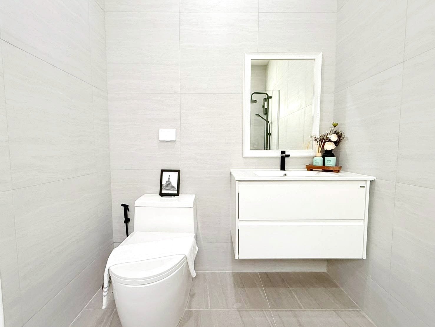 Modern white bathroom with a wall-mounted vanity, large mirror, and toilet; decorative toiletries on the counter.