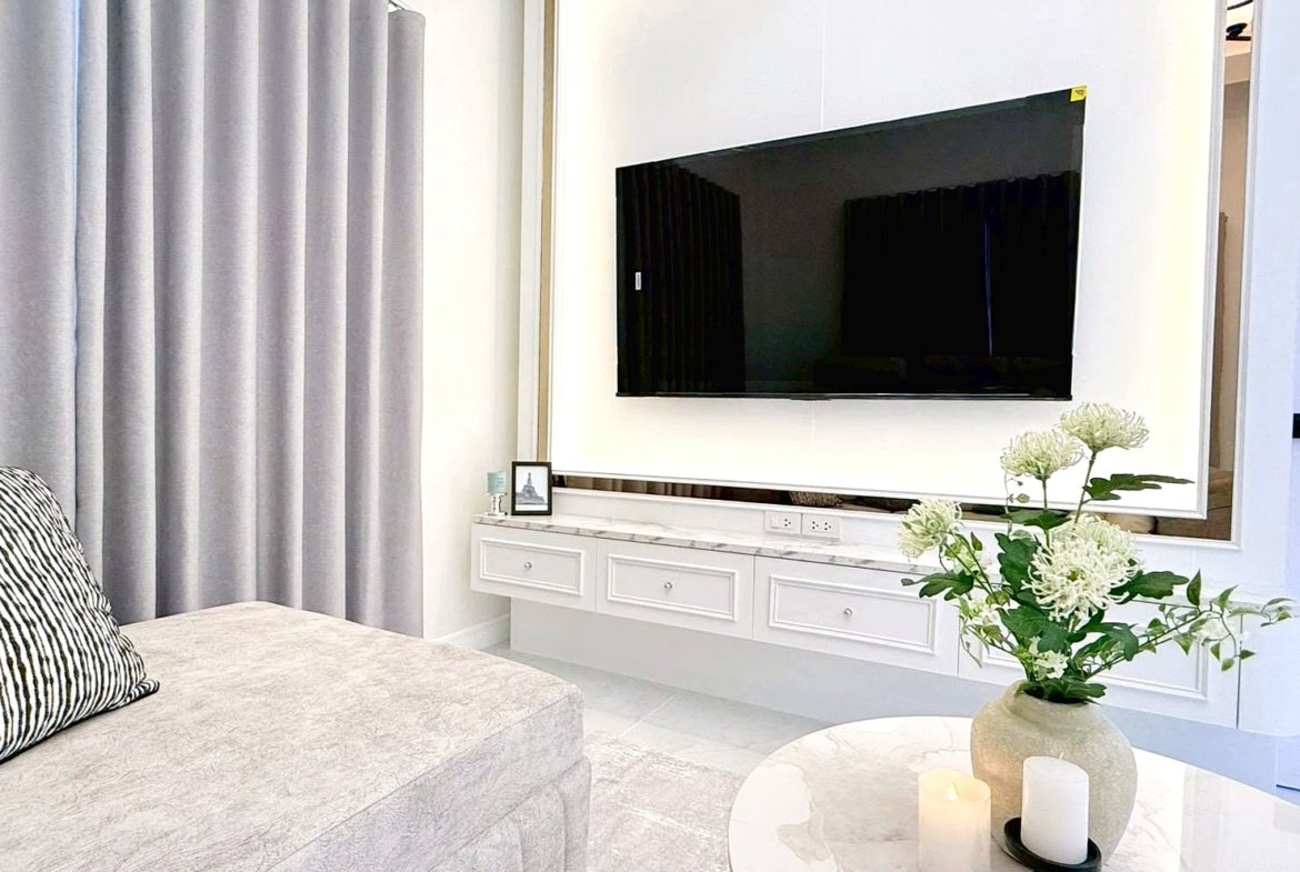 Modern living room with a large flat-screen TV mounted on a white wall above a marble console and a vase of white flowers on a round marble coffee table.