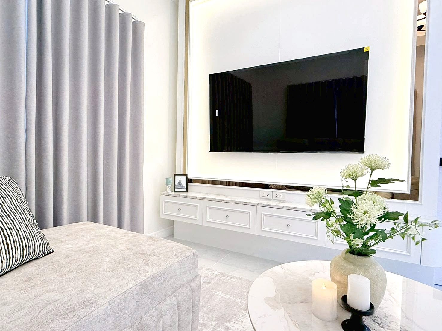 Modern living room with a large flat-screen TV mounted on a white wall above a marble console and a vase of white flowers on a round marble coffee table.