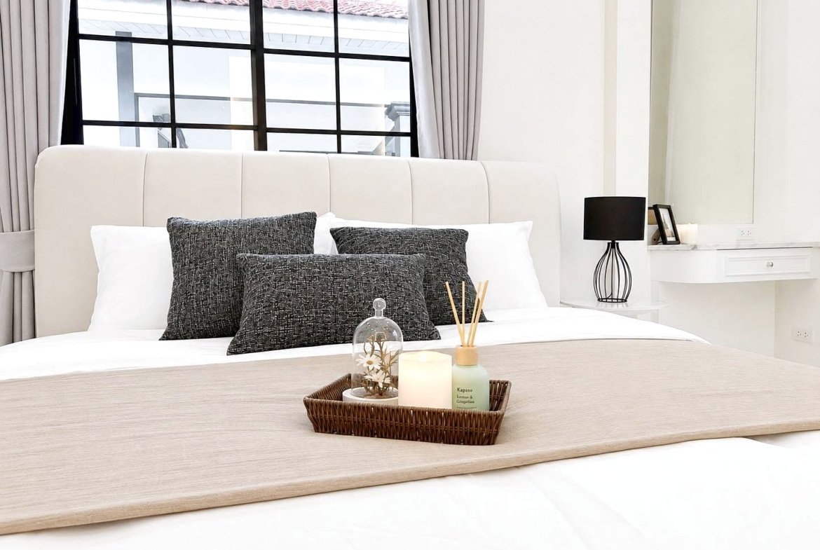 Neutral-toned bedroom with a white upholstered bed, dark gray pillows, and a beige runner; a wicker tray holds a candle and diffuser on the duvet.